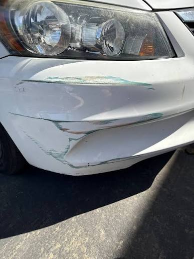 Green paint transfer smeared across the white front bumper of a car, below the headlight. The mark sits on top of the paint rather than cut into it.
